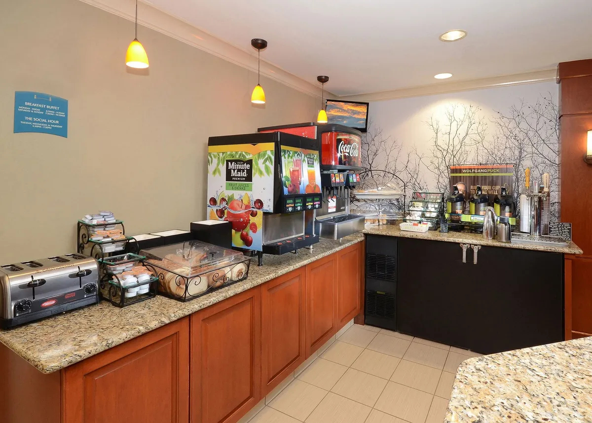 A bright, casual breakfast area featuring a well-stocked buffet counter with beverage dispensers, toasters, and a tempting pastry display. Guests gather to enjoy breakfast and mingle in a welcoming ambiance.
