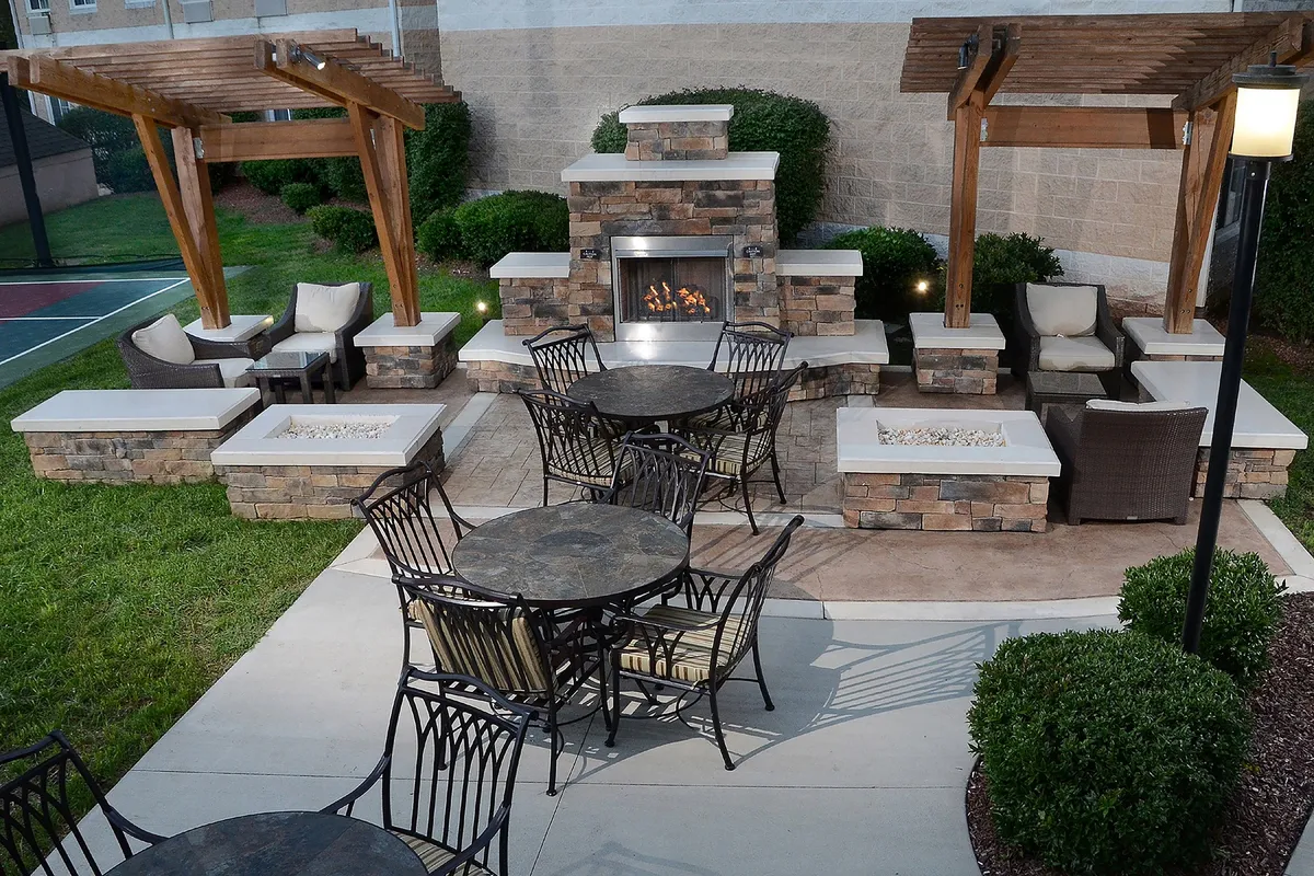 This exterior image showcases an inviting outdoor seating area centered around a striking fireplace, with tables and comfortable chairs surrounded by lush landscaping. The scene suggests a welcoming space for guests to relax, hold informal meetings, or enjoy social gatherings after a productive day.