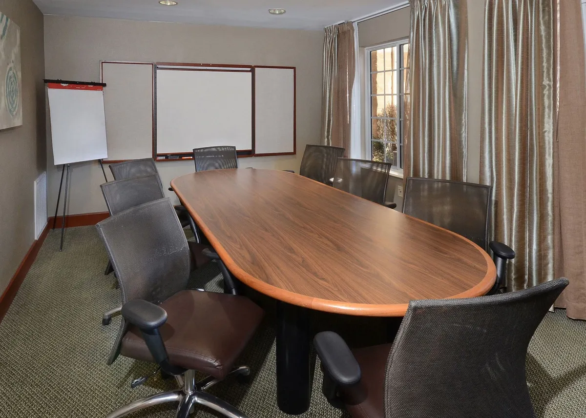 A modern business center with a large wooden conference table, comfortable office chairs, a presentation board, and natural light streaming through the window, offering an ideal environment for work or meetings.