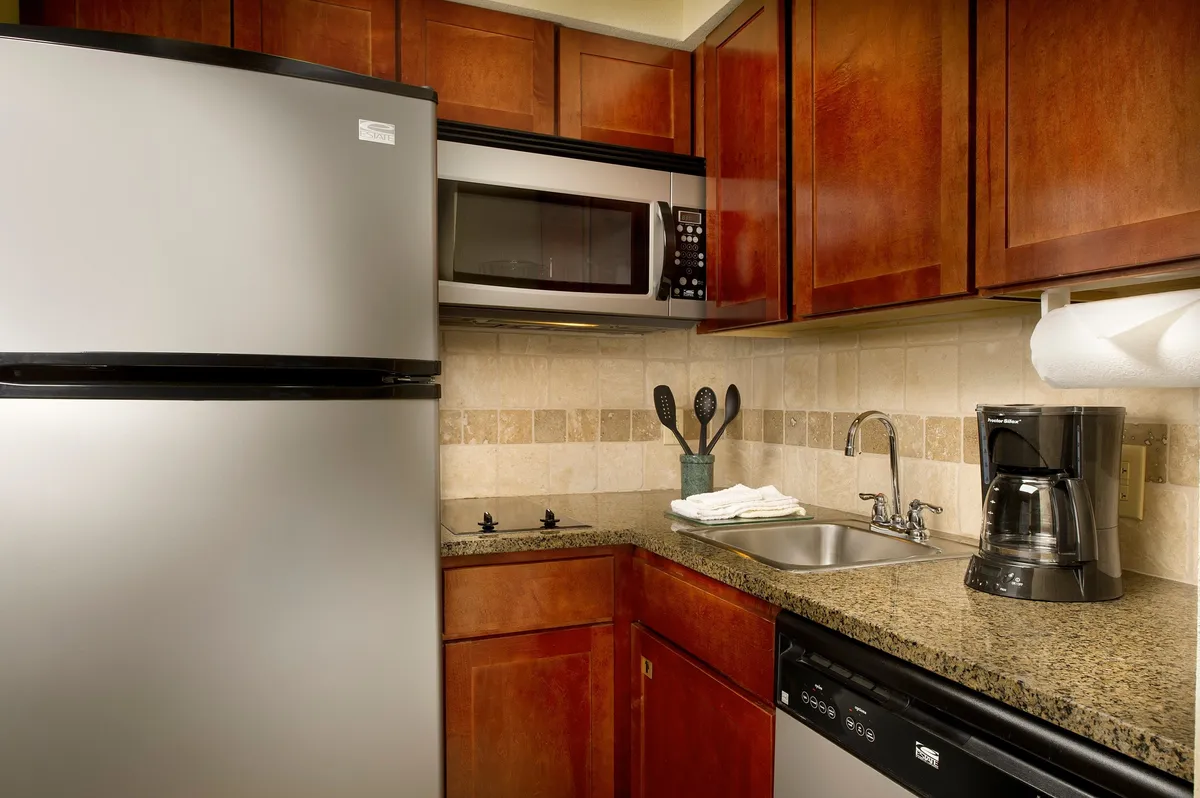 This image features a fully equipped kitchen with a silver refrigerator, microwave, coffee maker, sink with faucet, and granite countertop. The kitchen is practical and homey, capturing morning routines like brewing coffee and preparing meals.