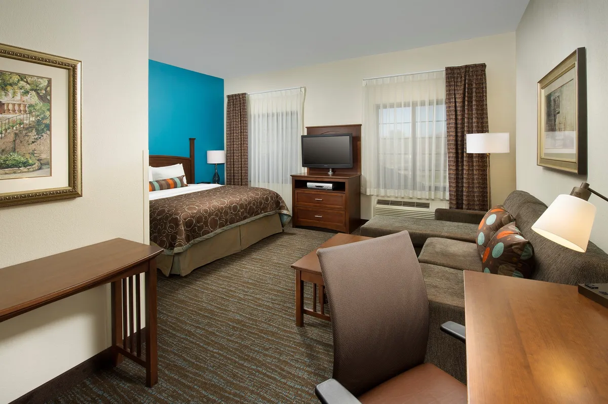 The image showcases a bright, thoughtfully arranged studio space featuring a plush queen bed, cozy sleeper sofa, dedicated work desk, and a flat-screen TV. Soft lighting, modern décor, and inviting artwork create a warm ambiance perfect for both relaxation and productivity. The scene exudes comfort and versatility, highlighting spaces where guests can enjoy morning coffee, unwind with a movie, or catch up on work.